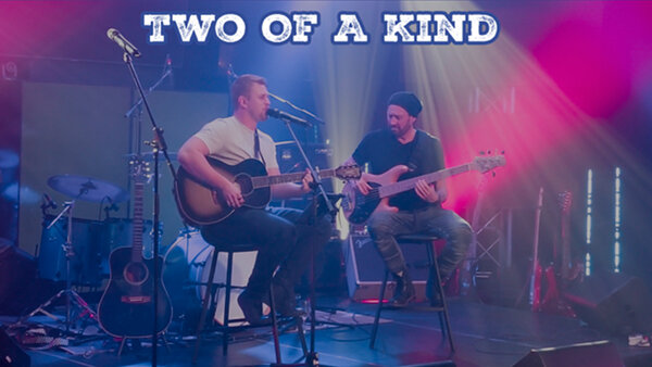Performance musicale du duo Two of a Kind à L'Hôtel Plaza, avec un guitariste et un bassiste sur scène sous des lumières colorées
