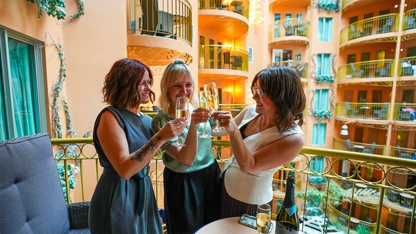 Trois femmes trinquent avec des coupes de champagne sur un balcon du Palace Royal, au cœur d’un atrium lumineux, pour une escapade dans le Vieux-Québec.