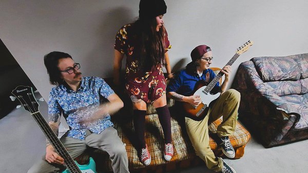 The Leftlovers en pleine vibe : un trio énergique sur un vieux divan, entre guitare électrique, basse turquoise et look rétro décontracté.