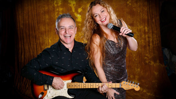 Duo Gaston et Monya lors d'une prestation musicale dans le décor chaleureux de l'Hôtel Québec Inn, mettant en scène Gaston jouant de la guitare et Monya chantant avec un micro.