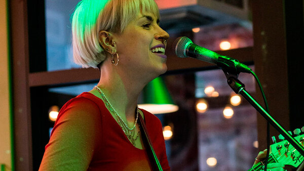 Cassandre Picard souriante, jouant de la guitare et chantant lors d'une prestation musicale à l'Hôtel Palace.