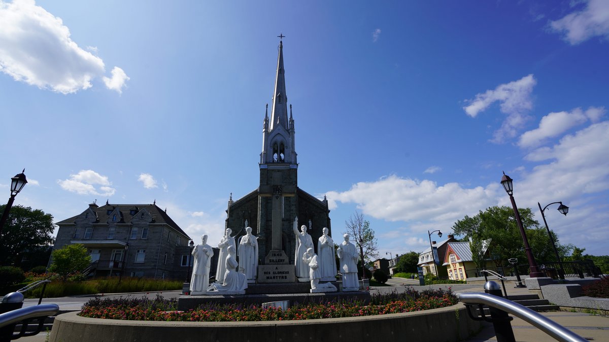 Site patrimonial de Sillery Église Saint-Michel de Sillery, avec sa flèche élancée, entourée d'un massif de statues blanches et de fleurs colorées sous un ciel ensoleillé.