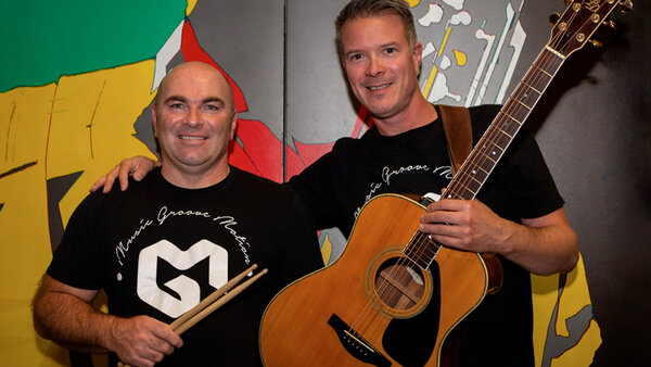 Duo musical MGM lors d'une prestation à l'Hôtel Palace, avec un musicien tenant une guitare acoustique et un autre avec des baguettes de batterie, devant un fond coloré.
