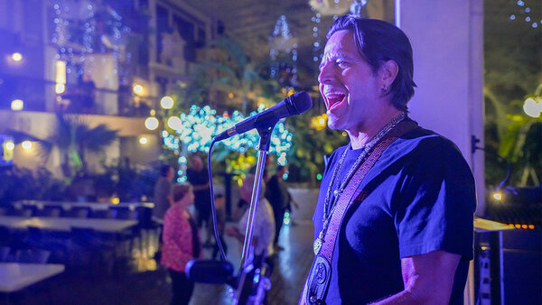 A musical presentation in a tropical garden at the Quebec Inn, featuring singer Remmy Langlois playing guitar, surrounded by a festive, illuminated atmosphere.