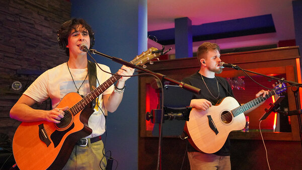 Musical performance in the Plaza Hotel by the duo The Lucky Ones. Two musicians play acoustic guitar while singing, with microphones and headphones.