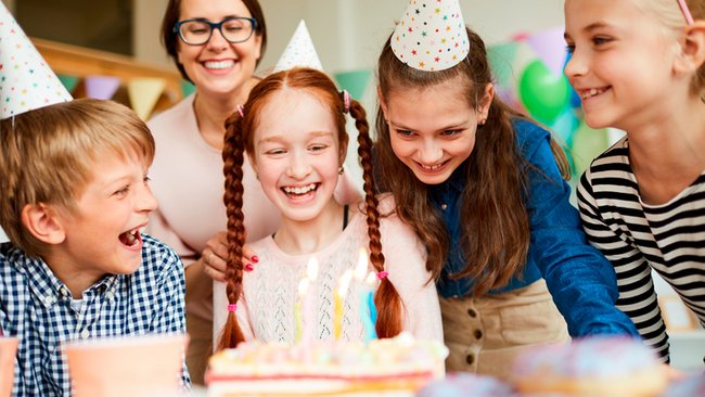 Un groupe d'enfants portant des chapeaux de fête sourient et rient autour d'un gâteau d'anniversaire avec des bougies allumées. Une femme en arrière-plan, également souriante, partage ce moment joyeux. Ambiance festive et colorée, idéale pour célébrer un anniversaire à L’Hôtel Québec avec le forfait spécial.
