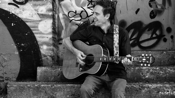Performance musicale de Louis Dionne à L'Hôtel Palace, jouant de la guitare acoustique dans un cadre urbain avec des graffitis en arrière-plan, créant une ambiance artistique et décontractée.