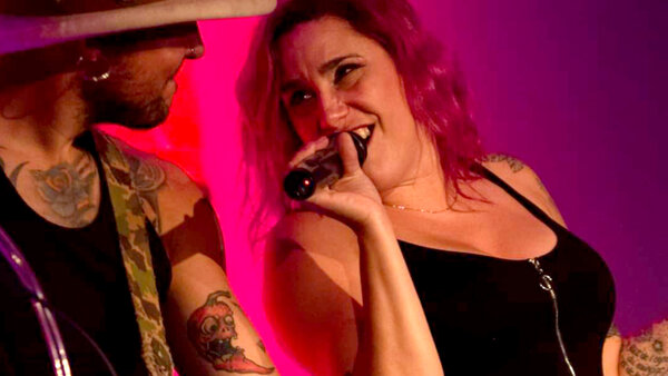 Cathy Vallières lors d'une prestation musicale à l'Hôtel Palace, chantant au micro avec énergie, accompagnée d'un guitariste sous un éclairage rose vibrant.